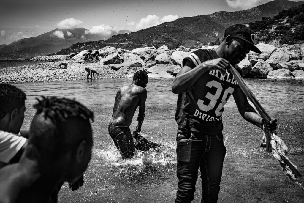 Sudanese boys wash their only clothes at the mouth of the Roja River, in front of beachgoers. After COVID-19, the Italian Red Cross camp in Ventimiglia was closed, leaving migrants without official support. 2020.