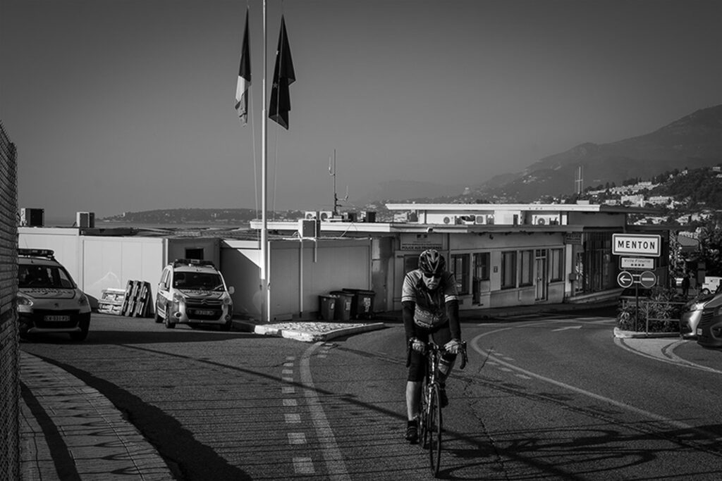An unaware cyclist crosses the border in front of the containers used by the PAF (Police aux Frontières) to detain migrants before their return to Italy. Since 2015, reports by organisations have documented cases of violence and abuse at the border.