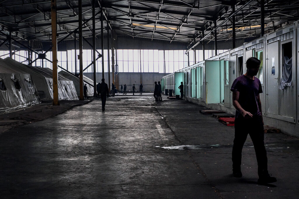 The IOM camp of Bira is located inside a former refrigerator factory. It offers around 2,000 people waiting to cross the border illegally a bed and a meal, nothing more. Tents, containers, and a space so vast that light fails to reach the people wandering through it, stripped of purpose and humanity. Bihać, Bosnia and Herzegovina, 2019.