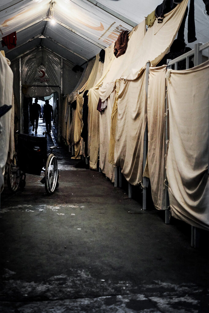 There are no windows in the camp and the walls are very high. All around, the fencing turns it into a prison. Many field tents bear the emblem of the Turkish Red Crescent. Each can host up to 150 people. Clothes and sheets are used to divide one sleeping space from another in a search for privacy. Bira refugees camp, Bihac, Bosnia Herzegovina, 2019.