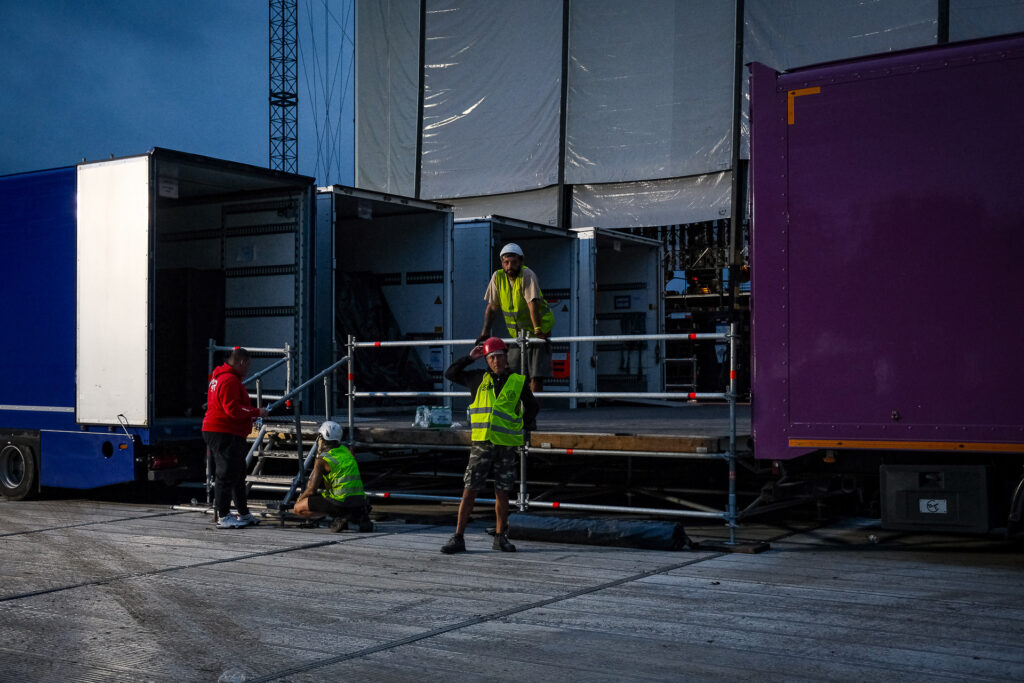 Andrea and Massimo are among the specialised stagehands called from across the country to work on the setup of a major concert stage, where hundreds of workers alternate day and night shifts while a large ramp behind the stage allows the simultaneous loading and unloading of up to eight trucks, part of a total of 47 vehicles involved in the production.