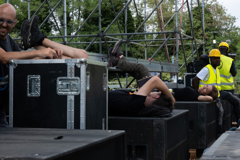 Technicians and stagehands rest on subwoofers and flight cases while waiting for more equipment to arrive before continuing the stage setup. Genoa, June 22nd 2022.