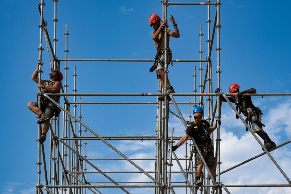 Rosario, Sebastiano, Bubacar and Michele, scaffholders specialised in stage construction, work on the dismantling of a concert stage. Like dancers of steel, their movements are fast and perfectly coordinated while the structure, no longer fully secured, starts swinging beneath their feet. Turin, July 2022.