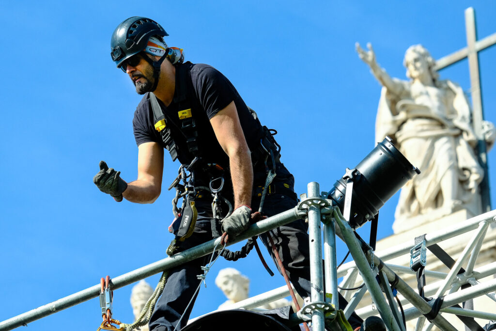 Roberto, a rigger, works high above Piazza San Giovanni in Rome. Constantly suspended at least ten metres above the ground, riggers are responsible for securing the audio and light systems to the truss structures suspended from the stage roof. Rome, April 2023