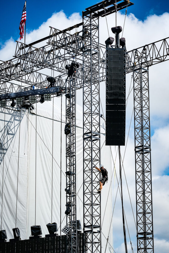 A rigger climbs the truss structure of a concert stage while securing the suspended line array speaker system high above the ground. Ferrara, August 2023