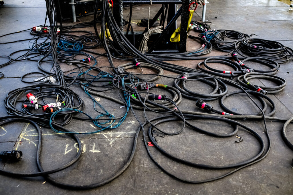 A tangled mass of audio, power and signal cables spreads across the stage floor during the setup of a live show. Behind the apparent chaos, every cable has a precise function and position within the technical system of the concert. Rome, April 2023.