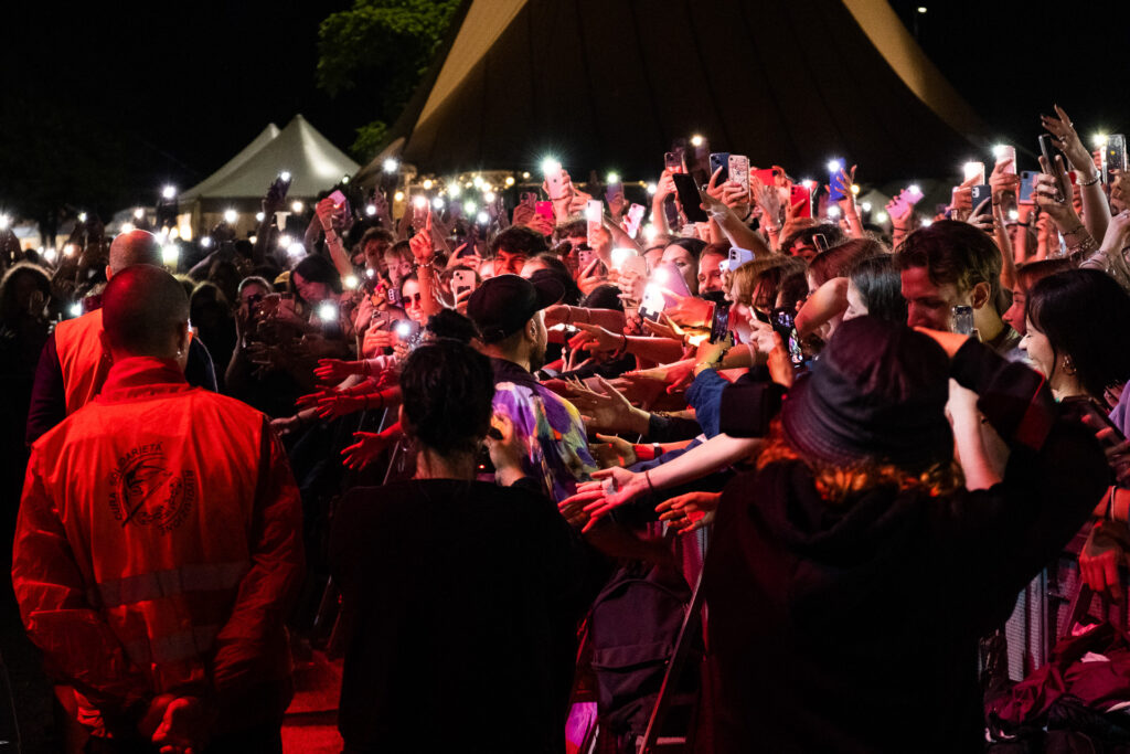 As the artist moves to the crowd, hundreds of hands and smartphones reach out in search of a brief moment of connection. Live concerts have increasingly become spaces where direct experience and digital mediation coexist, as audiences simultaneously seek to live, record and share the performance in real time. Padua, July 2022.