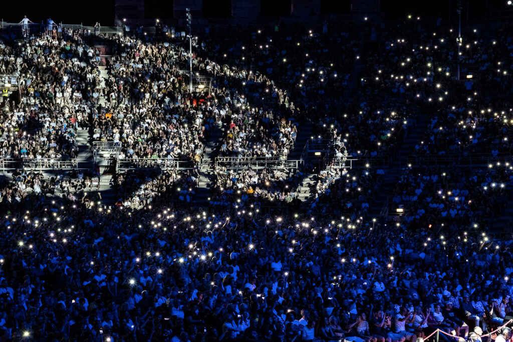 A shape of light illuminate the audience inside the Arena di Verona during a show. In a digital world, live music remains one of the few collective rituals capable of temporarily bringing strangers together within the same physical and emotional space. Arena di Verona, July 2022.