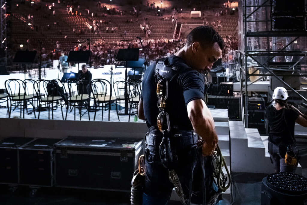 While the audience is still leaving the venue at the end of the show, Zack, a rigger, adjusts his harness before beginning the dismantling of the stage. Arena di Verona, July 2022.