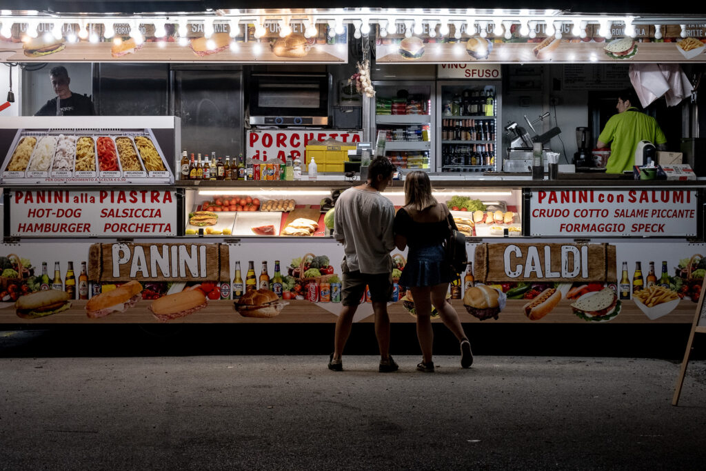 Outside the venue, a couple shares a late-night sandwich after the concert. Around live music revolves an entire temporary ecosystem of workers, vendors, relationships and small rituals that continue long after the lights on stage have gone out. Genoa, June 2022.