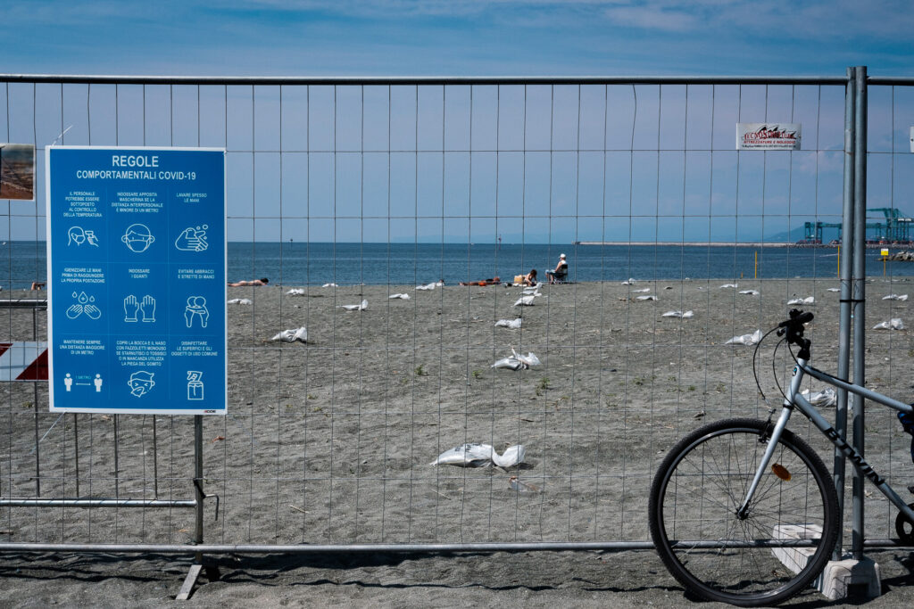 A sign at the entrance of the public beach in the working-class district of Voltri reminds visitors to wear masks, use hand sanitiser and maintain social distancing. Behind the fence surrounding the beach, the management of the Covid-19 emergency becomes visible through designated and separated spaces assigned to bathers. The beach, known locally as “La Fognazza” (“the sewer”), lies near oil terminals and the Fincantieri shipyards. The Port of Genoa is one of the most important maritime ports in Italy. Genoa, June 2nd 2020